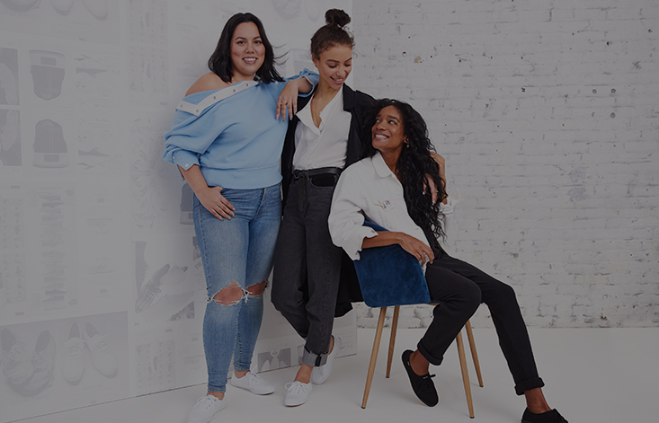 Three women pose against a white brick wall; two smile at each other