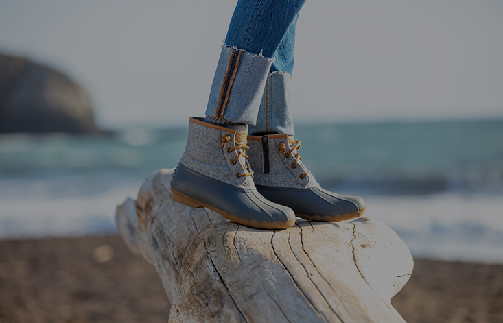 From the ankles down, a booted woman stands on a piece of wood near a coastline