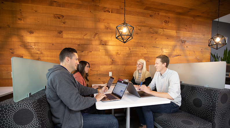 Two men and two women sit at a booth talking, with their laptops open