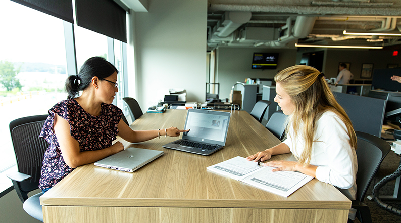 Two women sit opposite each other on a table while discussing something on a laptop