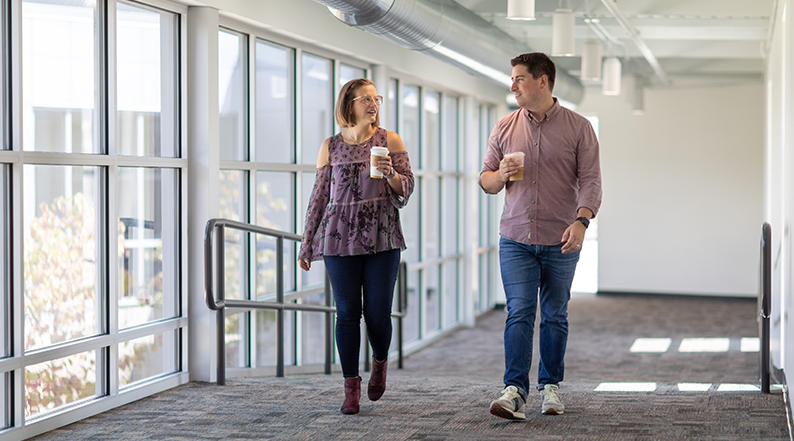 A man and a woman hold coffee cups as they walk together down a hallway