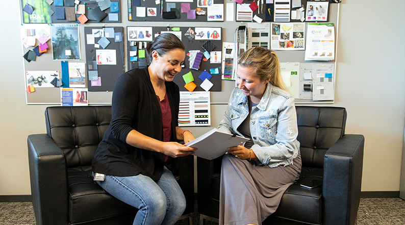 Two women talk, sitting in armchairs with product designs on the walls behind them