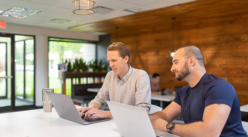 Two men talk at a table with their laptops open