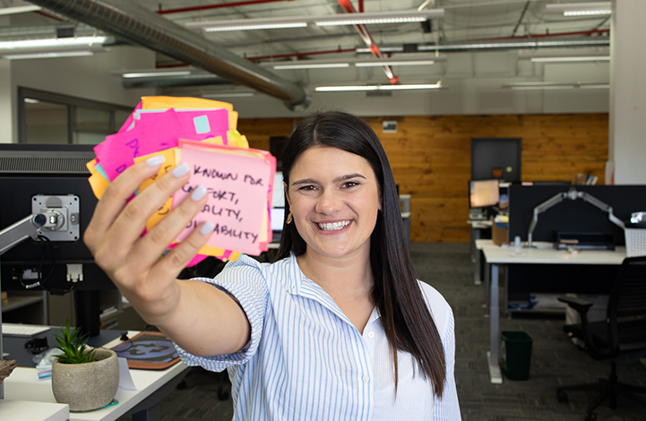 A woman smiles at the camera holding a stack of Post-it Notes