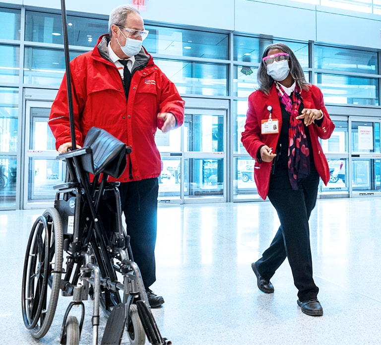 Male and female staff members wearing red jackets. The man is pushing an empty wheelchair