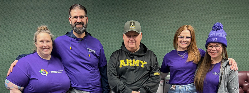 Four people with purple shirts stand on either side of a person wearing an Army hat and sweatshirt.