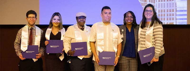 Six CNA program participants pose with their certificates before a large PowerPoint screen.