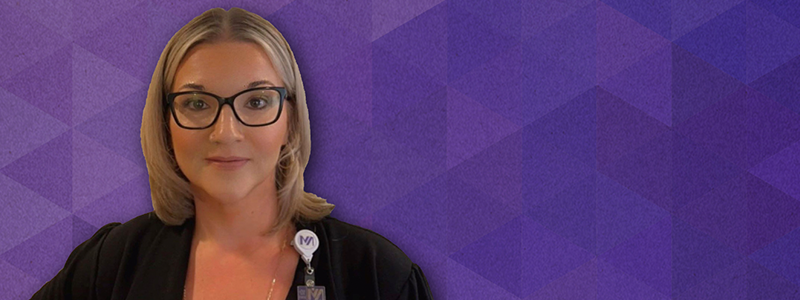 A white woman with blonde hair and black glasses softly smiles. Behind is a purple geometric background. 