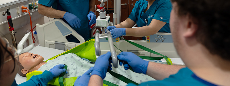 Four healthcare workers put a mannequin patient in a hydraulic lift. 