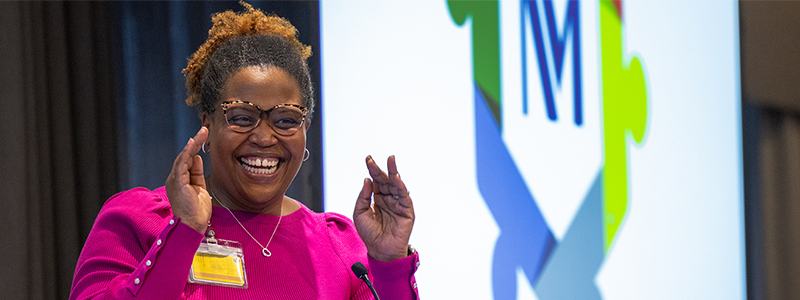 Khalilah Gates, MD, smiles while delivering a speech in front of a podium. She has dark brown skin and is wearing glasses and a bright pink sweater and skirt.