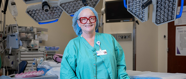 Portrait of Marianne Sheridan, RN, CNOR, in a surgery room wearing mint green scrubs and a blue hair covering.