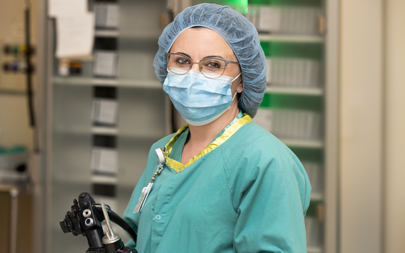Edibena Mussa poses on the job. She has fair skin. She wears glasses, a blue face mask, blue hair covering and seafoam-colored scrubs. She holds a black endoscope in her hands. 