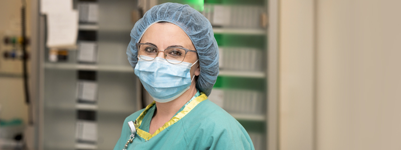 Edibena Mussa poses on the job. She wears glasses, a blue surgical mask, blue hair covering and green scrubs. She holds a black endoscope in her hands. 