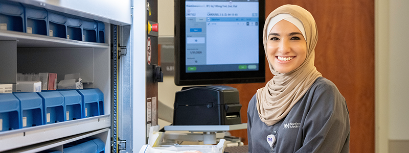 A person wearing hijab standing next to a counter with a pill bottle and hand scanner with computer behind them poses for a photo. 