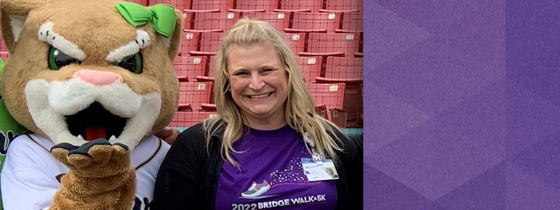 Tara Carlson wearing a purple t-shirt that says 2022 Bridge Walk 5k, standing next to a cougar baseball mascot who is blowing a kiss.