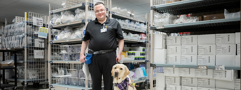 Christian Sullivan smiles while standing next to Zeke, his service dog. Christian wears a black shirt and pants and Zeke wears a purple, checkered bandana. 