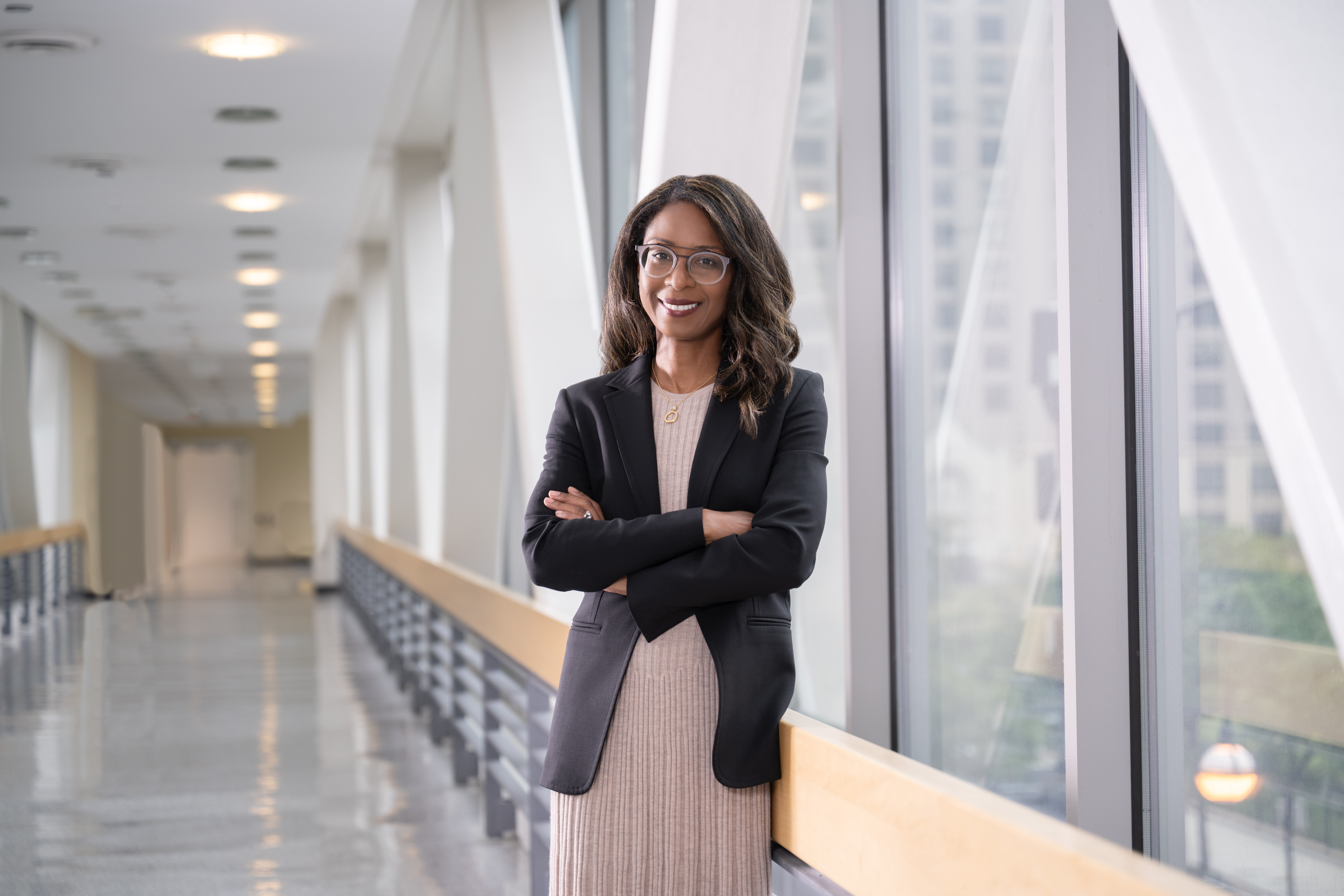 A Black woman stands and smiles in a hospital hallway. She is wearing a tan dress with a black jacket on top. She has glasses and shoulder-length brown hair.