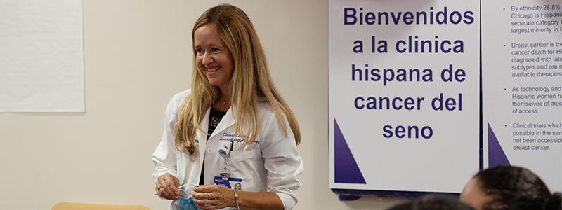 A physician with blonde hair and light skin smiles while holding a face mask. Behind her is a large sign in Spanish welcoming people to a breast cancer clinic. 