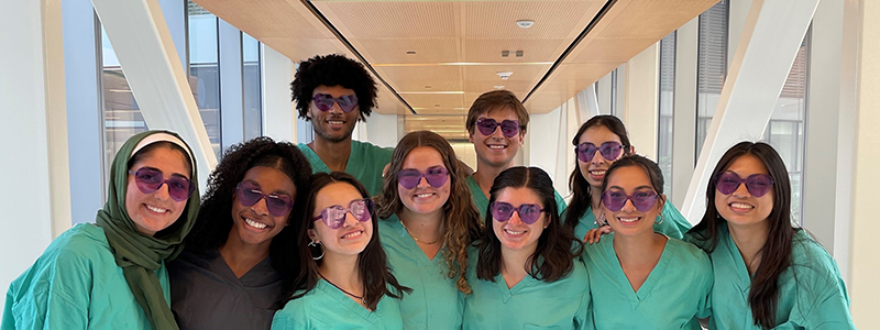 Twelve young adults wearing purple sunglasses and a mix of clinical and business attire pose for a photo in the skywalk.