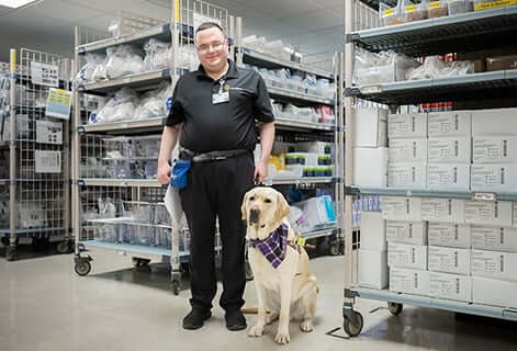 Christian Sullivan smiles while standing next to Zeke, his service dog. Christian wears a black shirt and pants and Zeke wears a purple, checkered bandana.