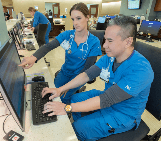 Two employees wearing scrubs look at a computer. 