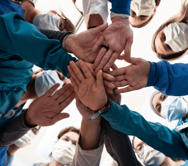 A group of employees wearing scrubs stands in a circle and stacks their hands together in the center.