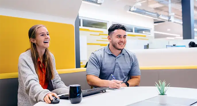 Two smiling students collaborate at a table in a modern office or study space.