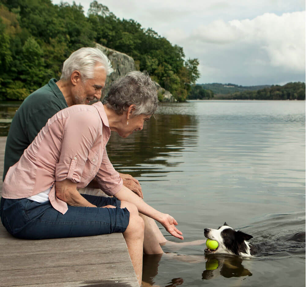 Older couple sitting on a dock and playing fetch with a swimming dog