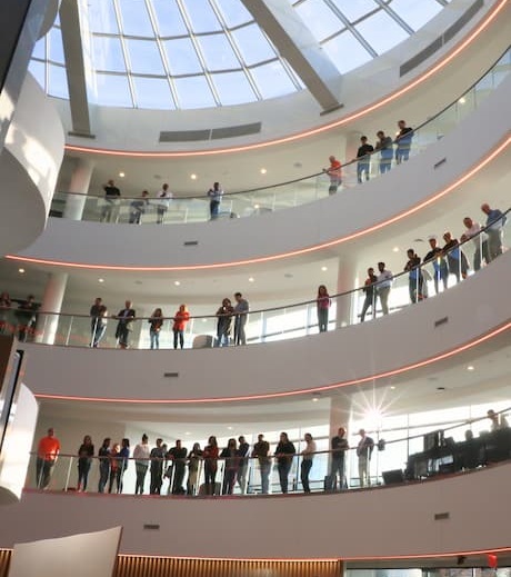 Large gathering of a crowd of workers in an open office with a balcony structure over a creative-looking set of work tables