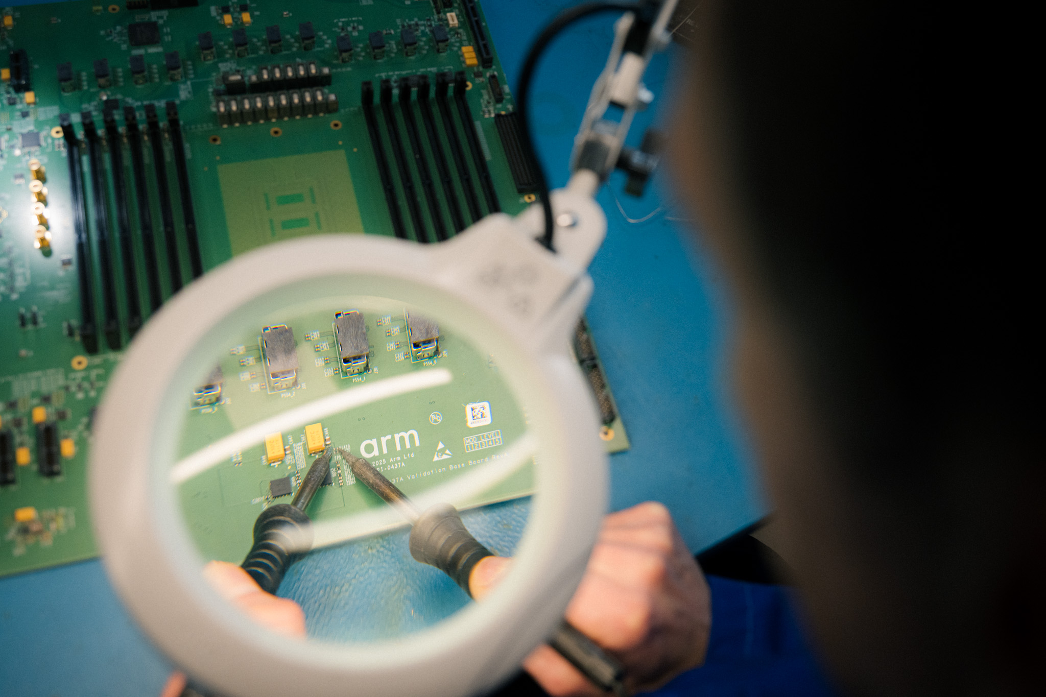 Engineer using a magnifying lamp and soldering tools to work on an Arm circuit board in a hardware lab.