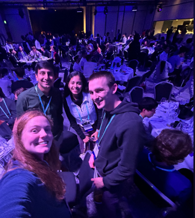 Four people smiling for a selfie at a large conference , with rows of attendees seated in an auditorium under blue and purple stage lighting.