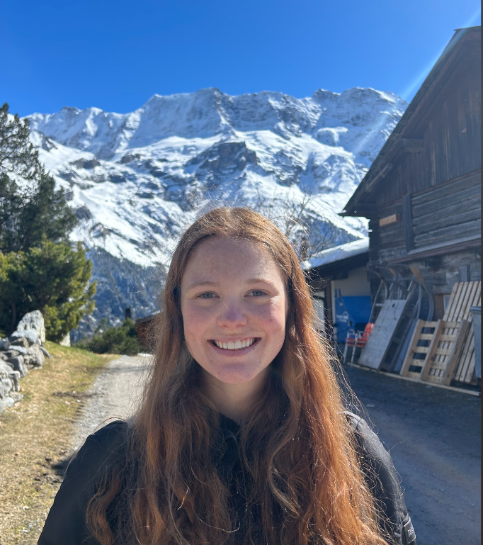 Audrey with long auburn hair smiling at the camera on a sunny mountain path, with snow-covered alpine peaks, evergreen trees, and traditional wooden chalets in the background under a clear blue sky.