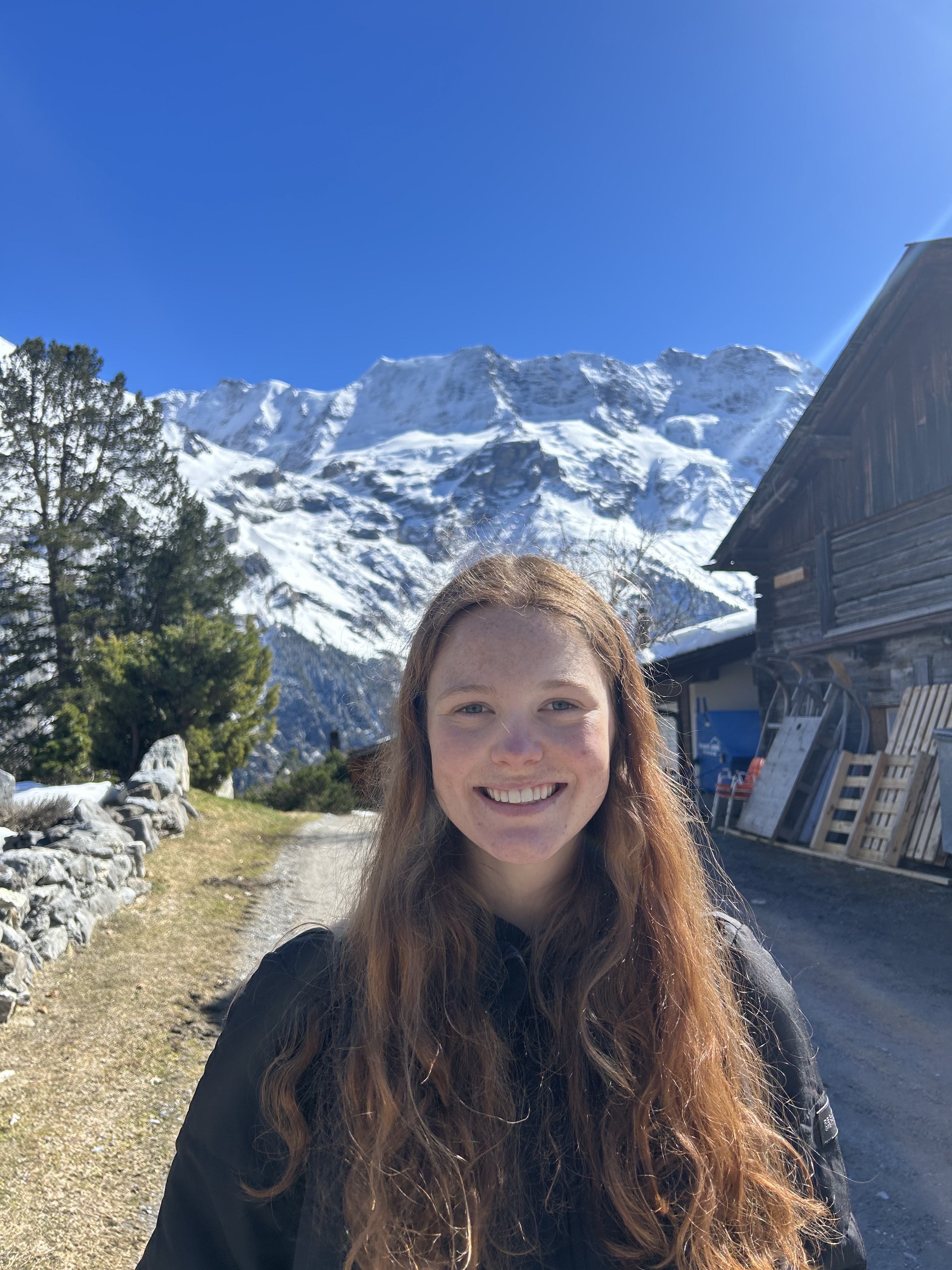Audrey with long auburn hair smiling at the camera on a sunny mountain path, with snow-covered alpine peaks, evergreen trees, and traditional wooden chalets in the background under a clear blue sky.