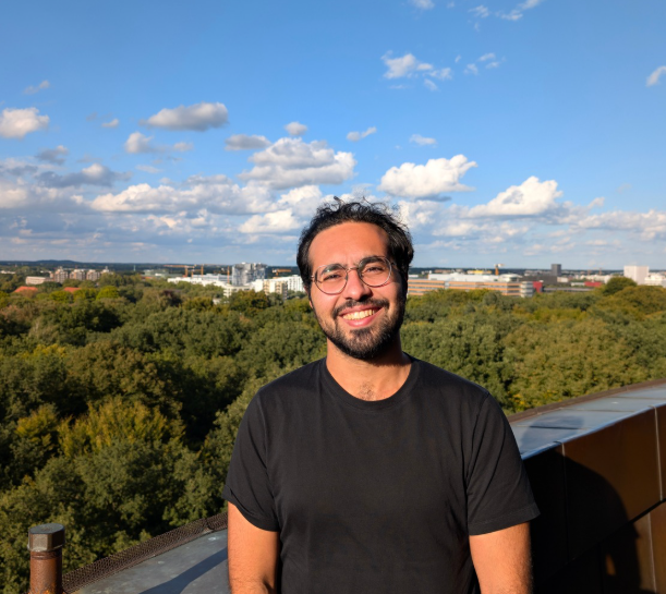 Omar smiling outdoors on a terrace with trees and a city skyline in the background.