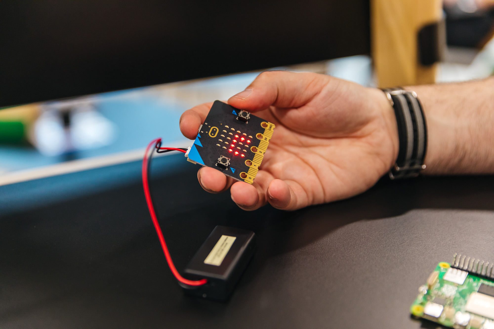 Person holding a microcontroller development board connected to a battery pack with wires on a desk.