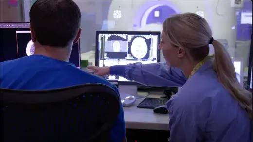 A female CT Technologist pointing to something on the screen while a male sits in a chair next to her (Video)
