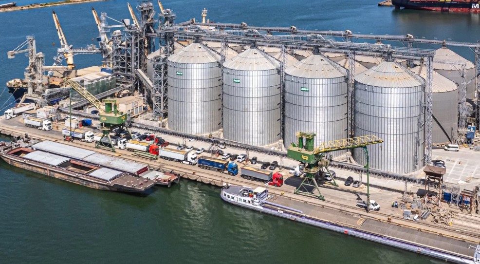 View of large silver grain silos at a port facility, with trucks, machinery, and water in the background