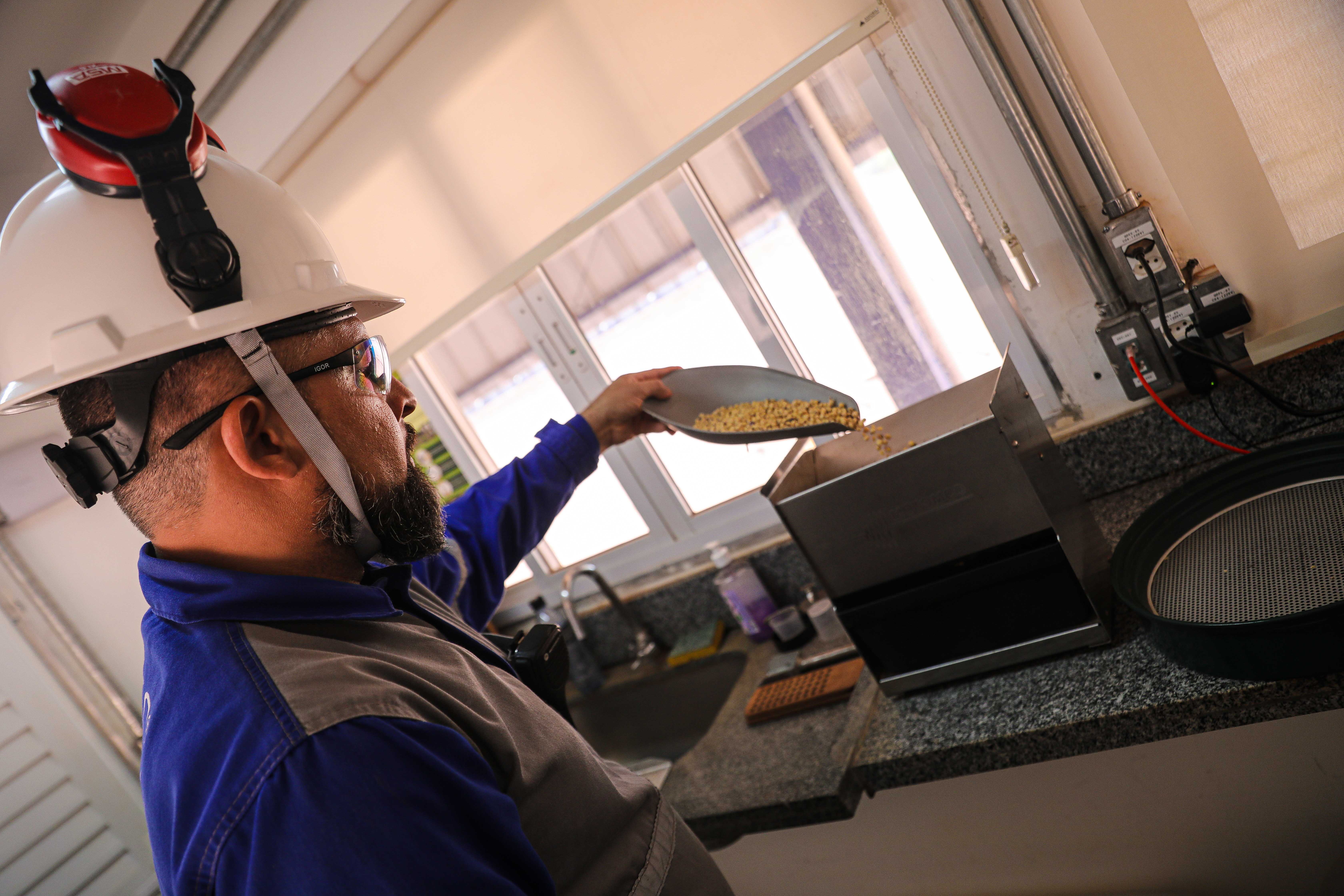 Trabalhador usando capacete branco e óculos de proteção despeja grãos de soja em uma máquina de teste dentro de um laboratório de controle de qualidade da CHS.