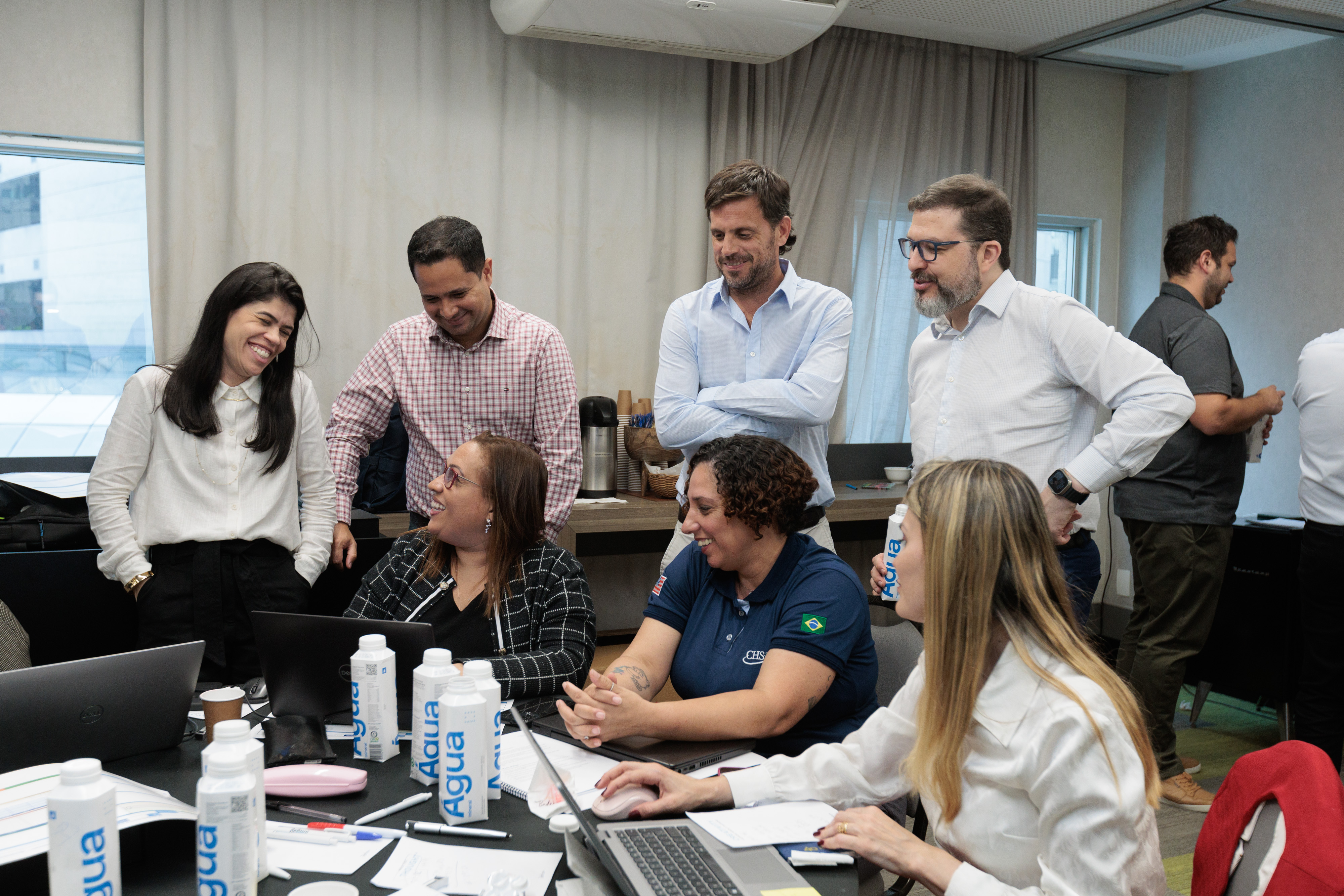 Grupo de profissionais colaborando em torno de uma mesa de conferência com laptops, cadernos e garrafas de água durante uma reunião de equipe em um ambiente de escritório.