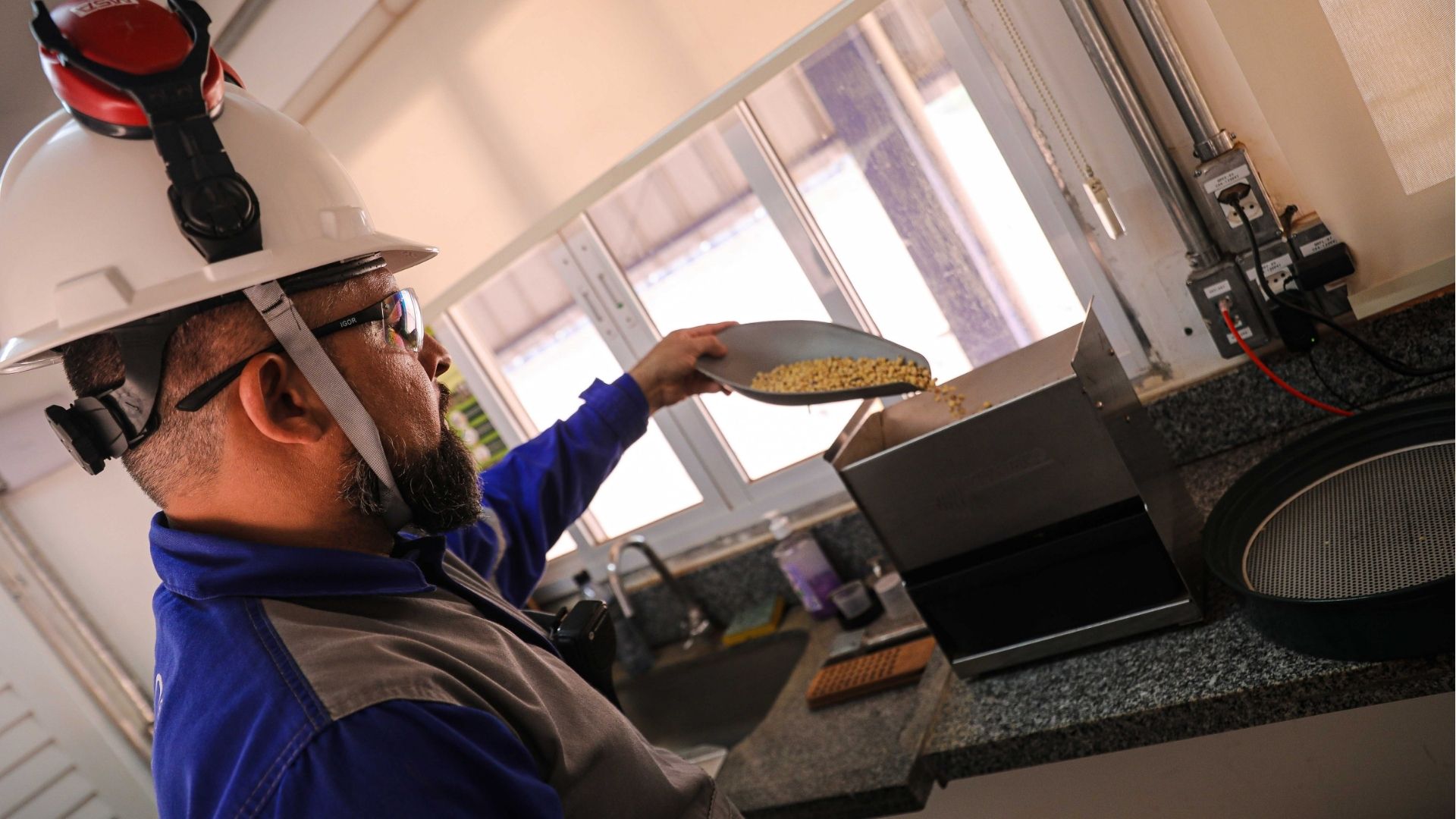 Trabalhador usando capacete branco e óculos de proteção despeja grãos de soja em uma máquina de teste dentro de um laboratório de controle de qualidade da CHS.