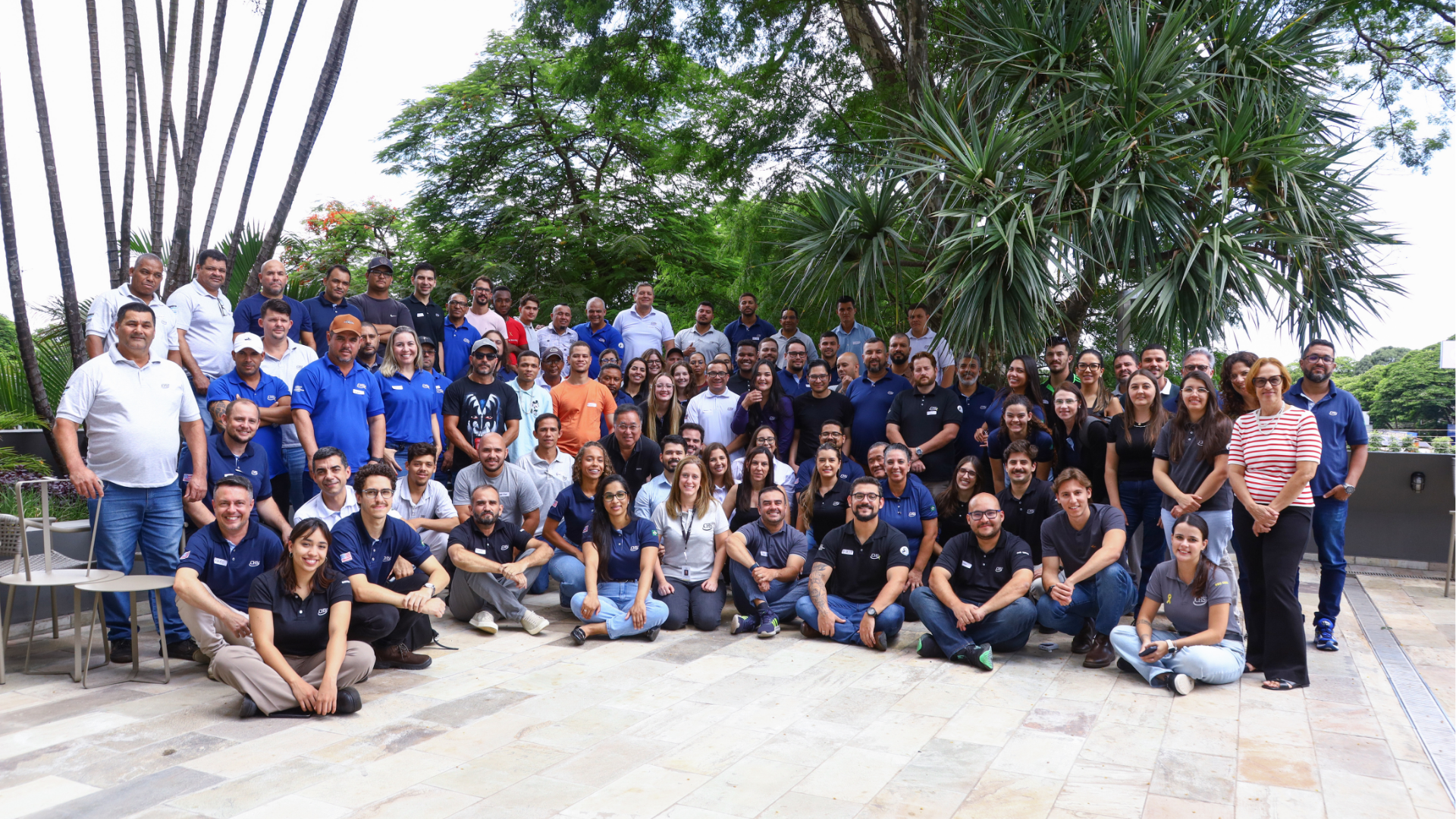 Gran grupo de empleados reunidos al aire libre, sonriendo y posando juntos en un patio ajardinado.