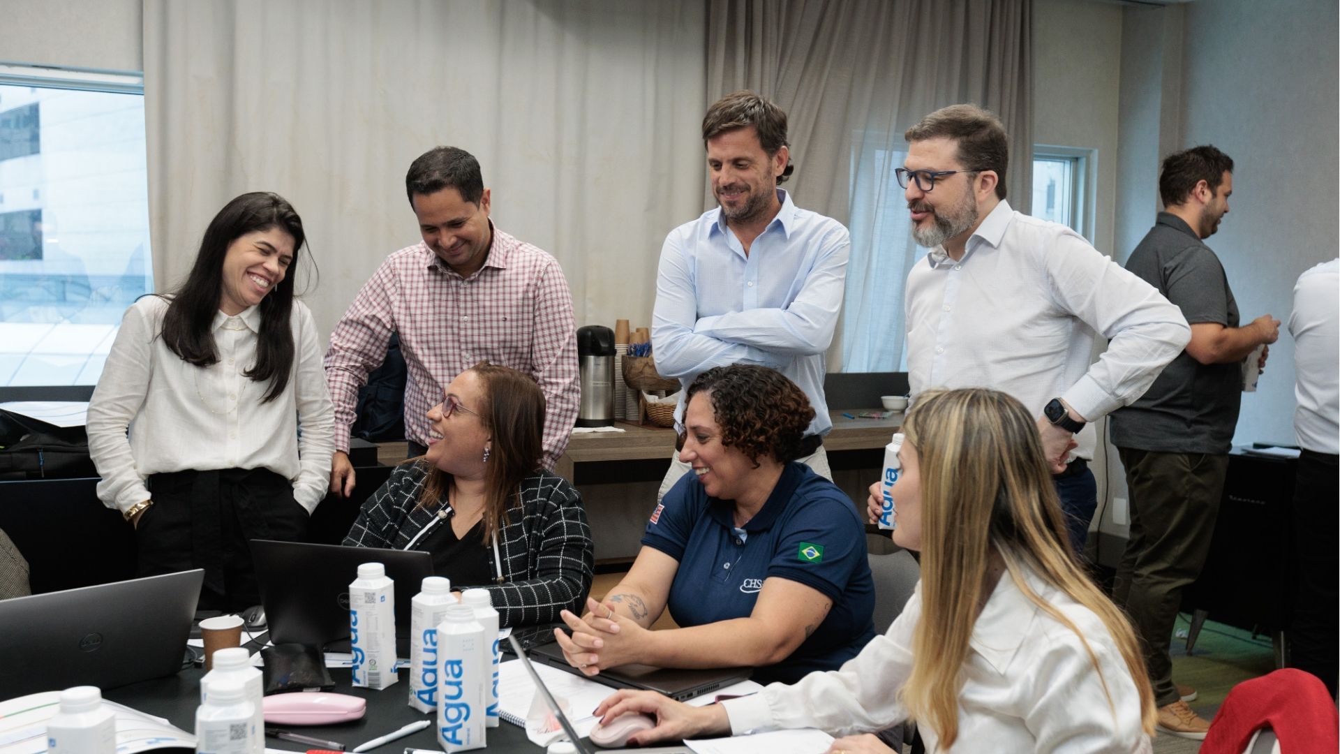 Grupo de profissionais colaborando em torno de uma mesa de conferência com laptops, cadernos e garrafas de água durante uma reunião de equipe em um ambiente de escritório.