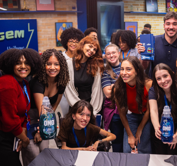 Grupo de pessoas reunidas em ambiente interno, posando próximo a uma mesa e segurando frascos de produto OMO, com elementos de marca Unilever ao fundo.