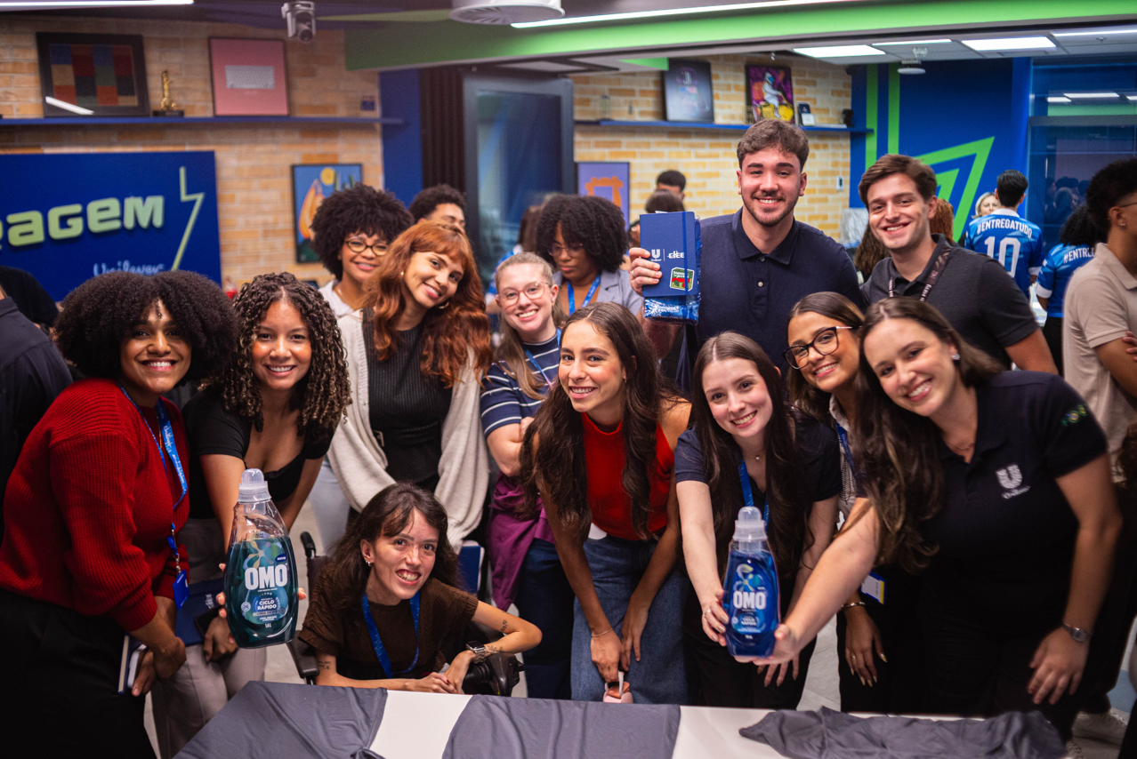 Grupo de pessoas reunidas em ambiente interno, posando próximo a uma mesa e segurando frascos de produto OMO, com elementos de marca Unilever ao fundo.