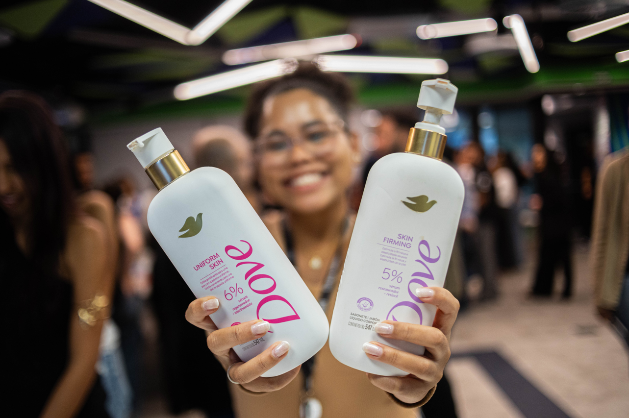 Pessoa em primeiro plano segurando dois frascos de produtos Dove diante da c&acirc;mera, em ambiente interno com pessoas e luzes ao fundo desfocadas.