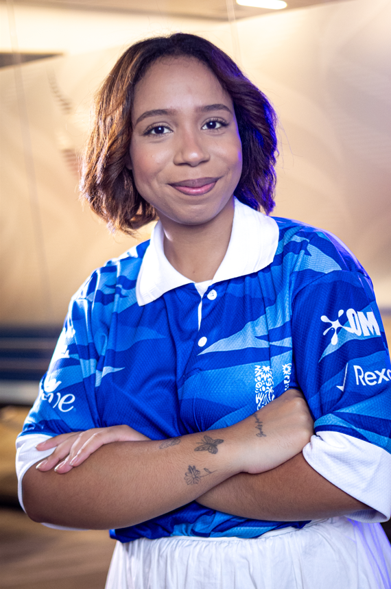 Garota negra sorridente em retrato de meio corpo, com braços cruzados, usando camisa azul esportiva da Unilever com colarinho branco, em ambiente interno iluminado.