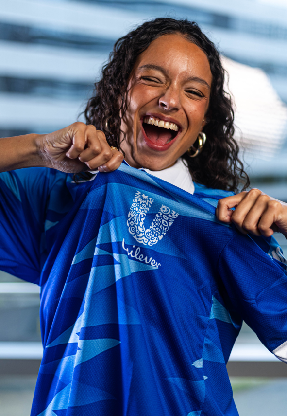Pessoa em close segurando a gola de uma camisa esportiva azul para destacar o logo da Unilever, com fundo urbano desfocado.