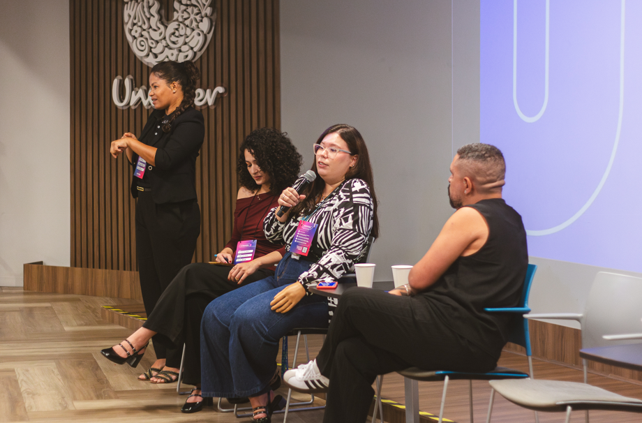 Grupo de quatro pessoas em painel da Unilever, sentadas no palco; uma fala ao microfone enquanto outra faz interpretação em libras ao lado.
