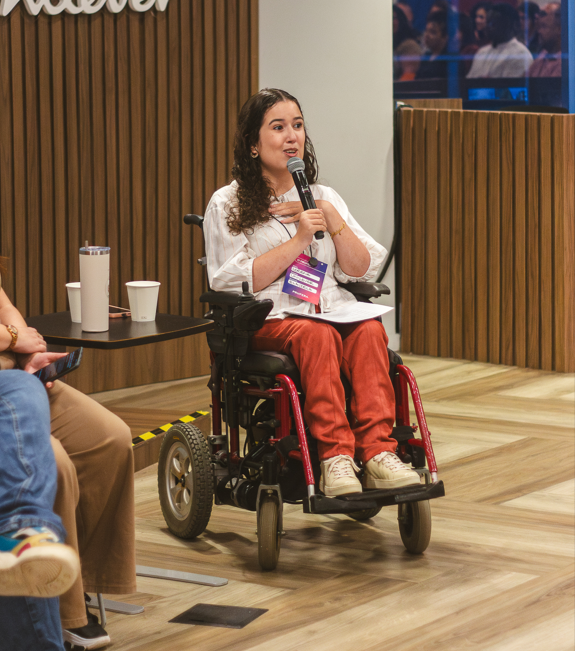Pessoa participante de painel corporativo fala ao microfone sentada em cadeira de rodas elétrica, em palco da Unilever.