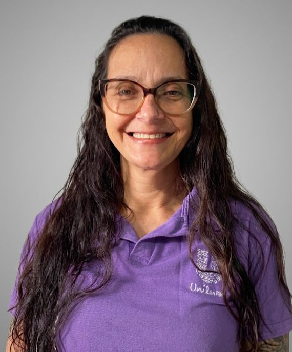 Jaqueline é uma mulher branca de cabelos castanhos longos. Está sorrindo e usa um óculos de armação marrom e uma camiseta polo na cor roxa, com o logo da Unilever